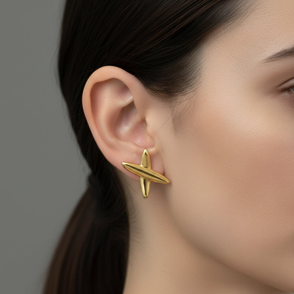 A woman with a sleek ponytail wearing large, 18K gold-plated, three-dimensional four-point cross-star stud earrings.