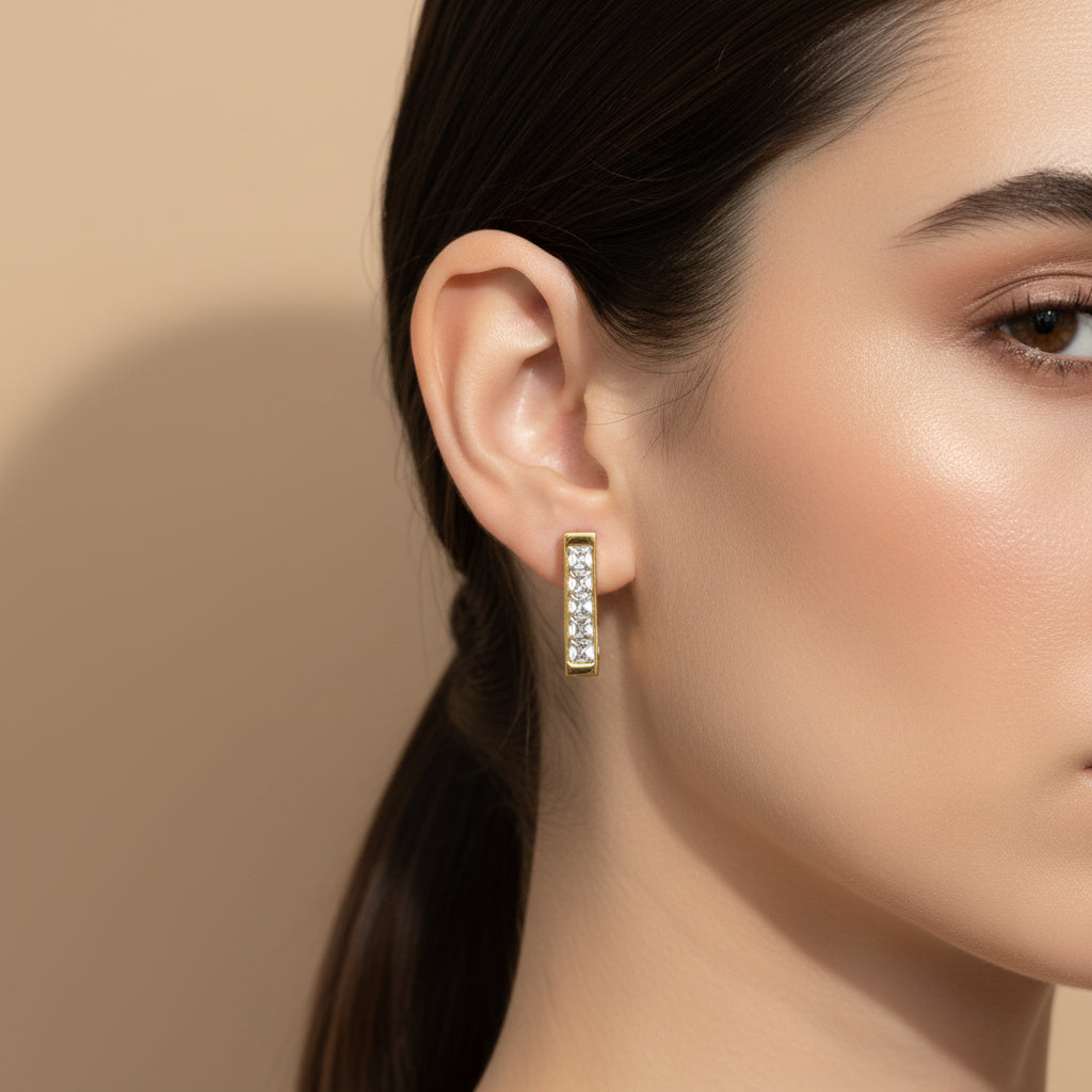 A woman with a sleek ponytail wearing 18K gold-plated vertical bar stud earrings featuring a row of clear baguette-cut crystals and a high-polish finish.