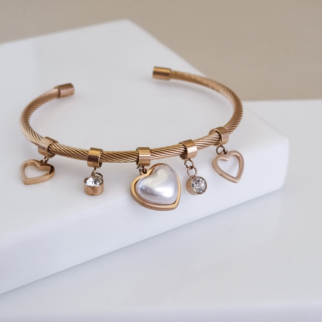 A high-polish 18K rose gold-plated open twisted cable cuff featuring heart and pearl charms, displayed on a minimalist white surface.