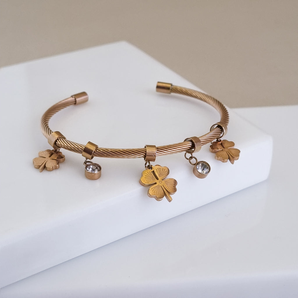 A high-polish 18K rose gold-plated open twisted cable cuff featuring sculpted four-leaf clover and crystal charms, displayed on a white surface.