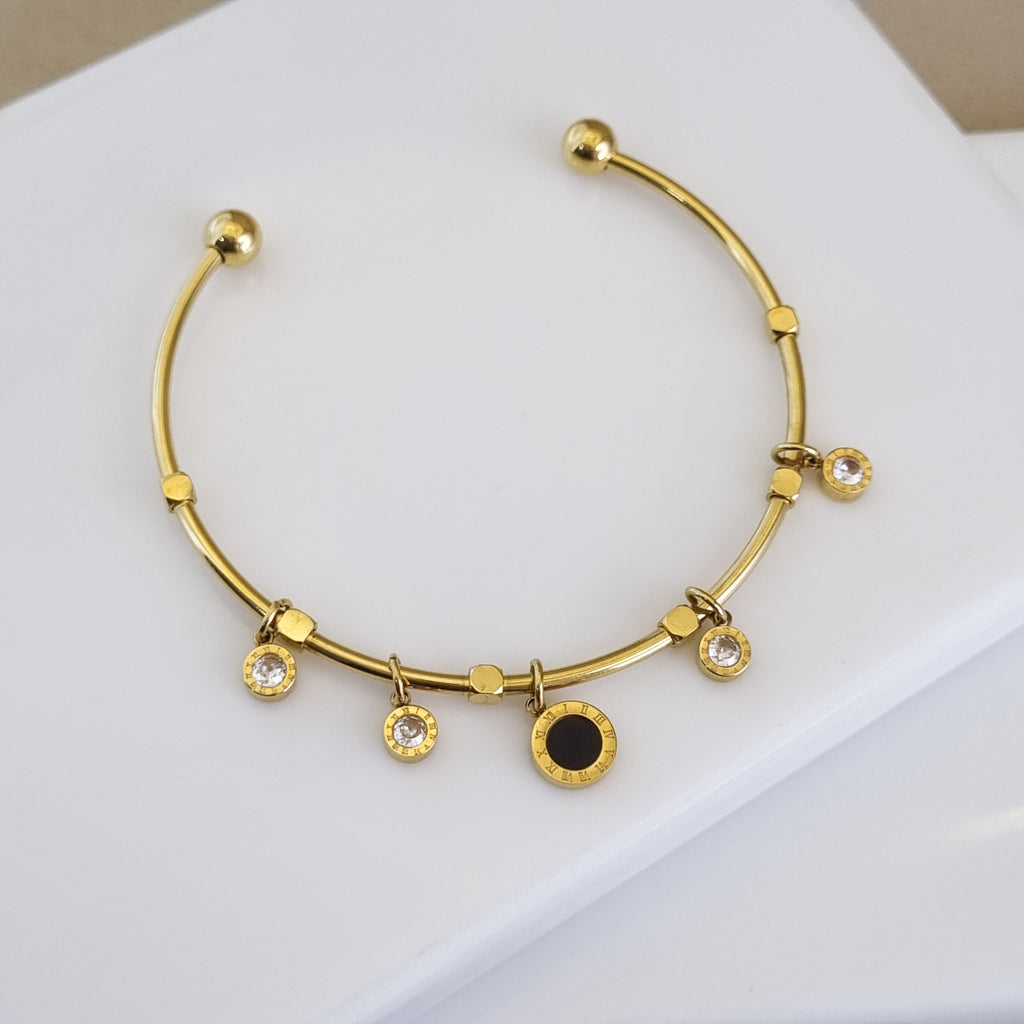 A high-polish 18K gold-plated open cuff bracelet featuring a central Roman numeral medallion and four bezel-set crystal charms on a white surface.