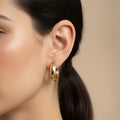 A woman with a sleek ponytail wearing medium-sized, 18K gold-plated thick tubular hoop earrings with a high-polish reflective finish.