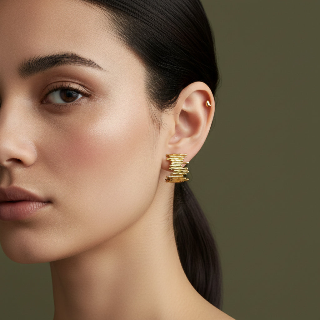 A woman with a sleek hairstyle wearing unique 18K gold-plated stud earrings featuring a horizontal stack of raw-edged golden bars.