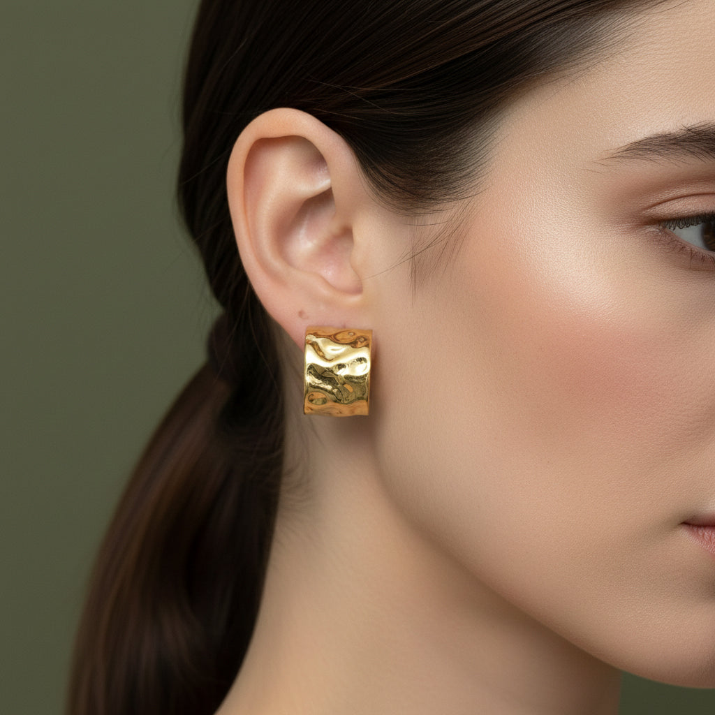 A woman with a sleek ponytail wearing large, 18K gold-plated wide cuff stud earrings featuring a deeply hammered high-polish texture.