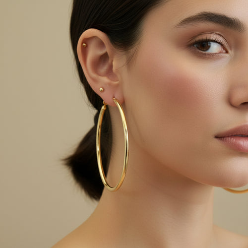 A woman with a sleek low bun wearing large, 18K gold-plated tubular hoop earrings with a high-polish reflective finish.
