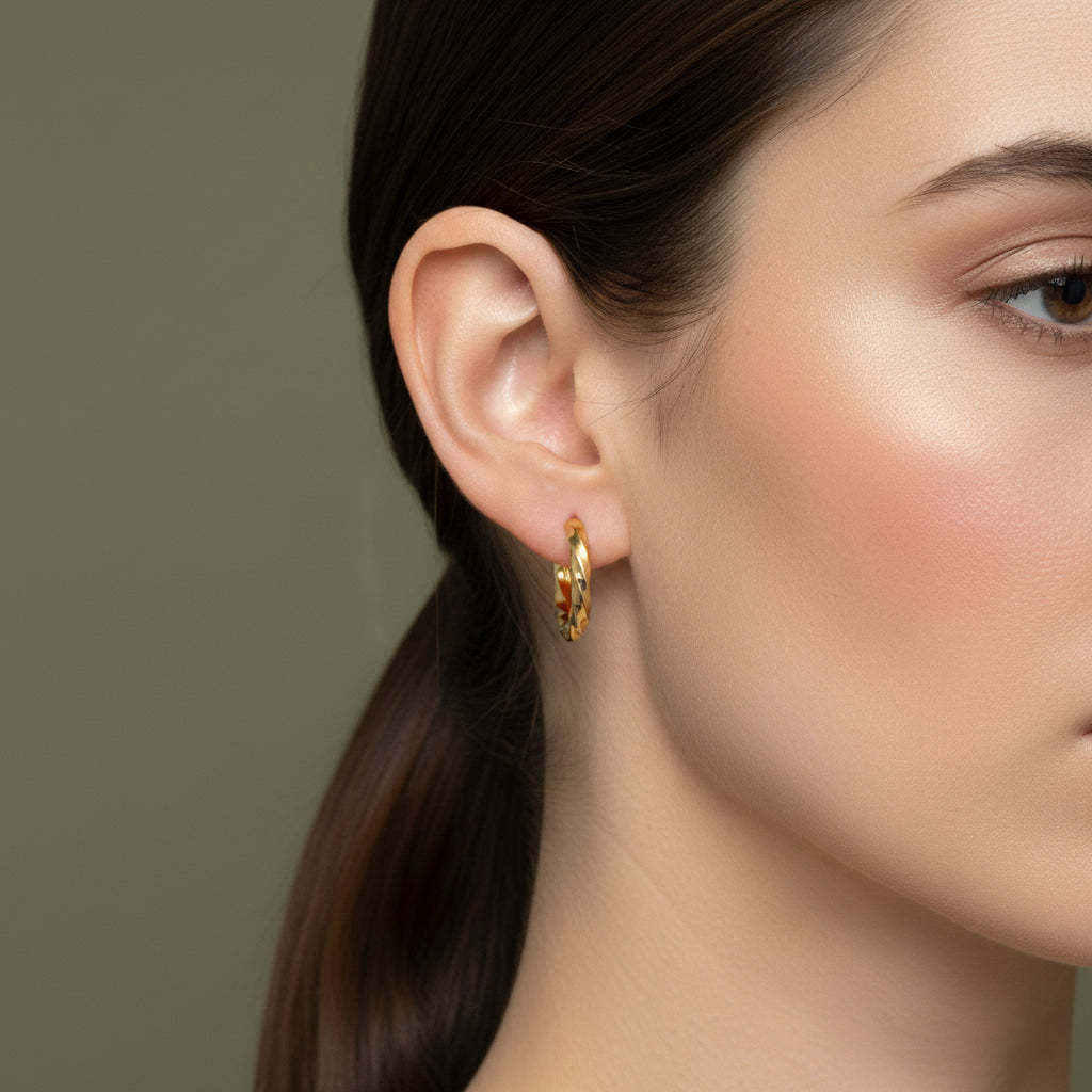 A woman with a sleek ponytail wearing small, 18K gold-plated huggie hoop earrings featuring a continuous helical twist and a high-polish finish.