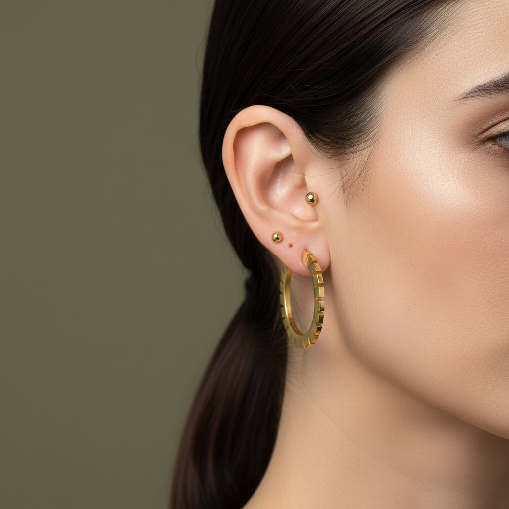 A woman with a sleek ponytail wearing large, 18K gold-plated flat-edge hoop earrings featuring precision-cut vertical notches and a high-polish finish.