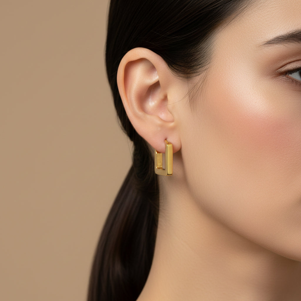 A woman with a sleek ponytail wearing small, 18K gold-plated square-shaped huggie hoop earrings with a high-polish reflective finish.