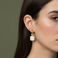 A woman with sleek dark hair wearing 18K gold-plated huggie hoop earrings with a large square iridescent drop and a high-polish finish.