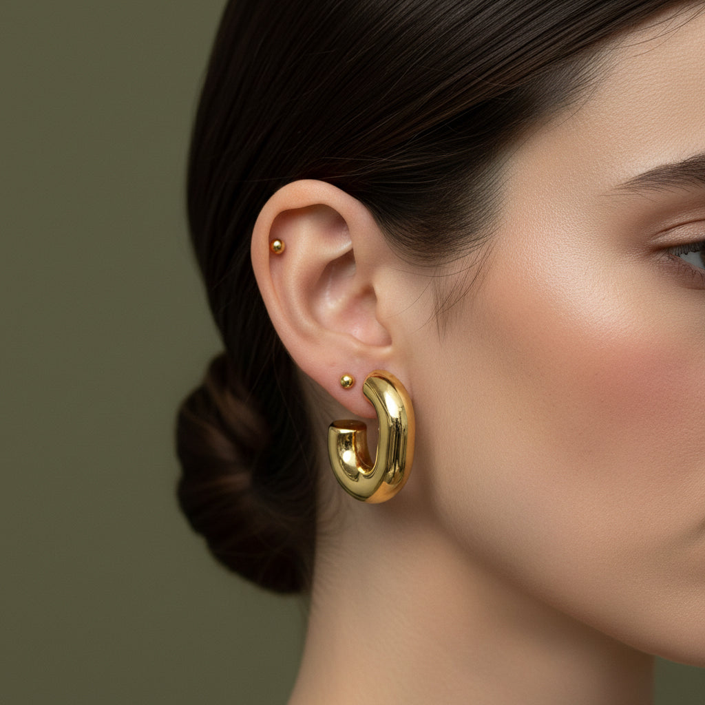 A woman with a sleek low bun wearing large, 18K gold-plated tubular "J" shaped hoop earrings with a high-polish reflective finish.