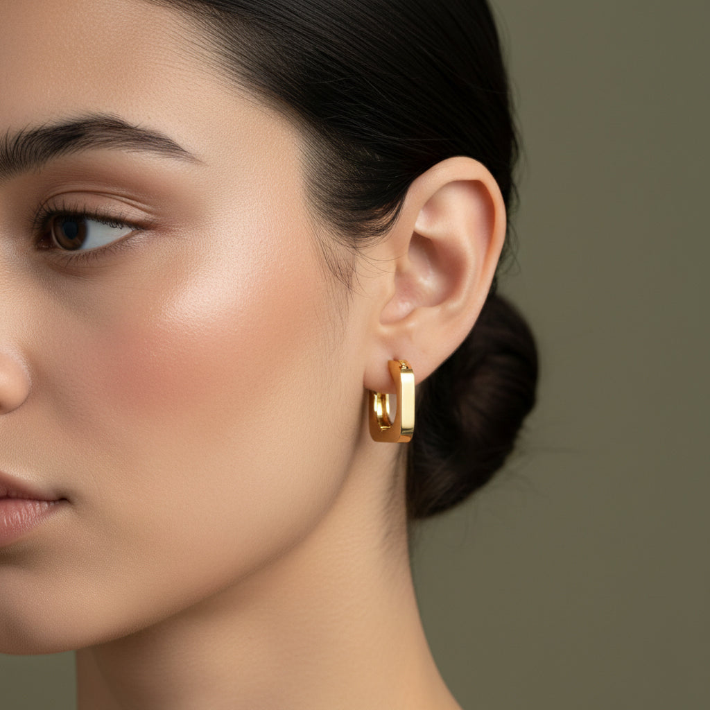 A woman with a sleek low bun wearing small, 18K gold-plated rectangular-shaped huggie hoop earrings with a high-polish reflective finish.