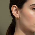 A woman with a sleek ponytail wearing small, thick 18K gold-plated micro tubular hoop earrings with a high-polish reflective finish.