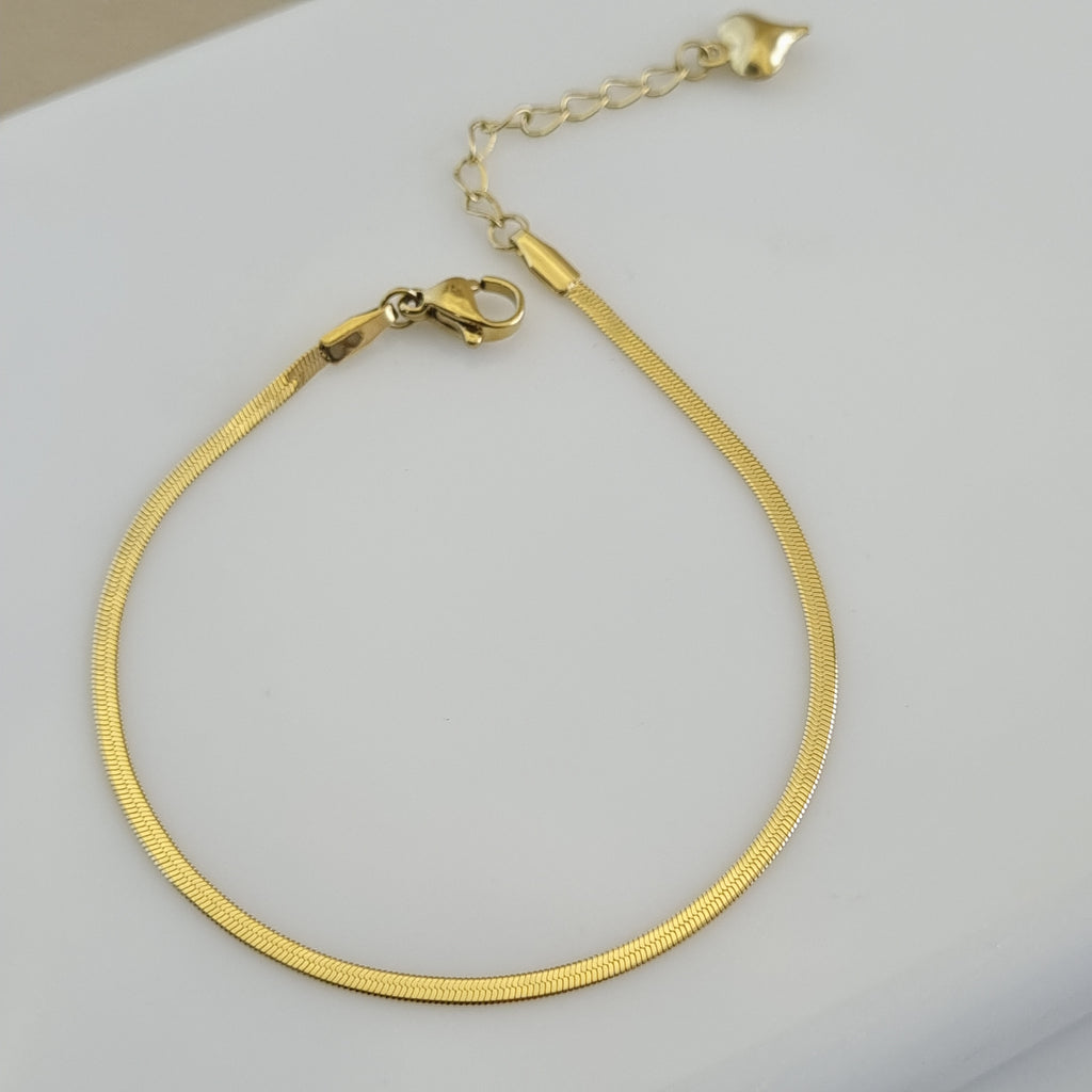 Treshire 18K gold plated Siena slim snake chain bracelet with a flat herringbone weave and heart tag on a white surface.