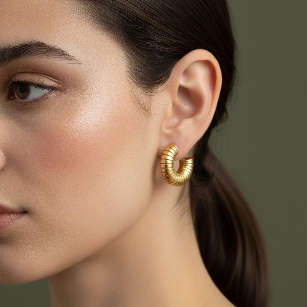 A woman with a sleek ponytail wearing 18K gold-plated "C" shaped hoop earrings featuring a continuous horizontal ribbed texture and high-polish finish.