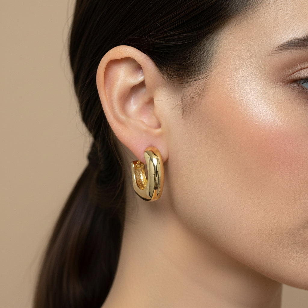 A woman with a sleek ponytail wearing 18K gold-plated thick "C" shaped huggie hoop earrings with a high-polish reflective finish.