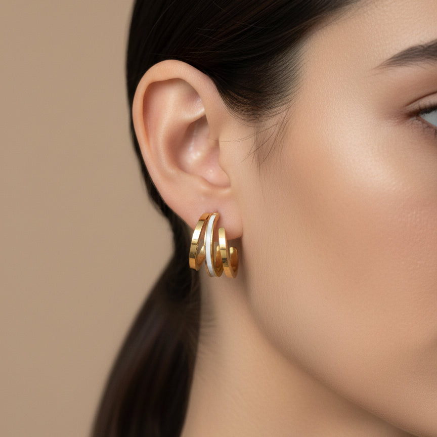 A woman with a sleek ponytail wearing 18K gold-plated triple-band huggie hoop earrings featuring a central white iridescent inlay and a high-polish finish.