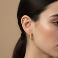 A woman with a sleek ponytail wearing small, 18K gold-plated square-shaped huggie hoop earrings with a high-polish reflective finish.