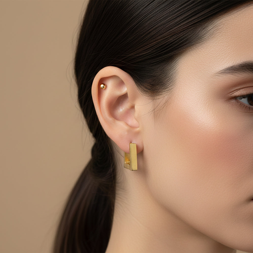 A woman with a sleek ponytail wearing small, 18K gold-plated square-shaped huggie hoop earrings with a high-polish reflective finish.