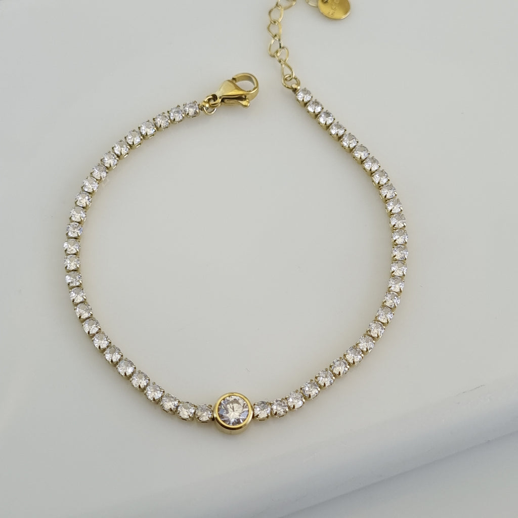 Treshire 18K gold plated tennis bracelet featuring a line of small crystals and a larger central bezel-set solitaire on a white surface.