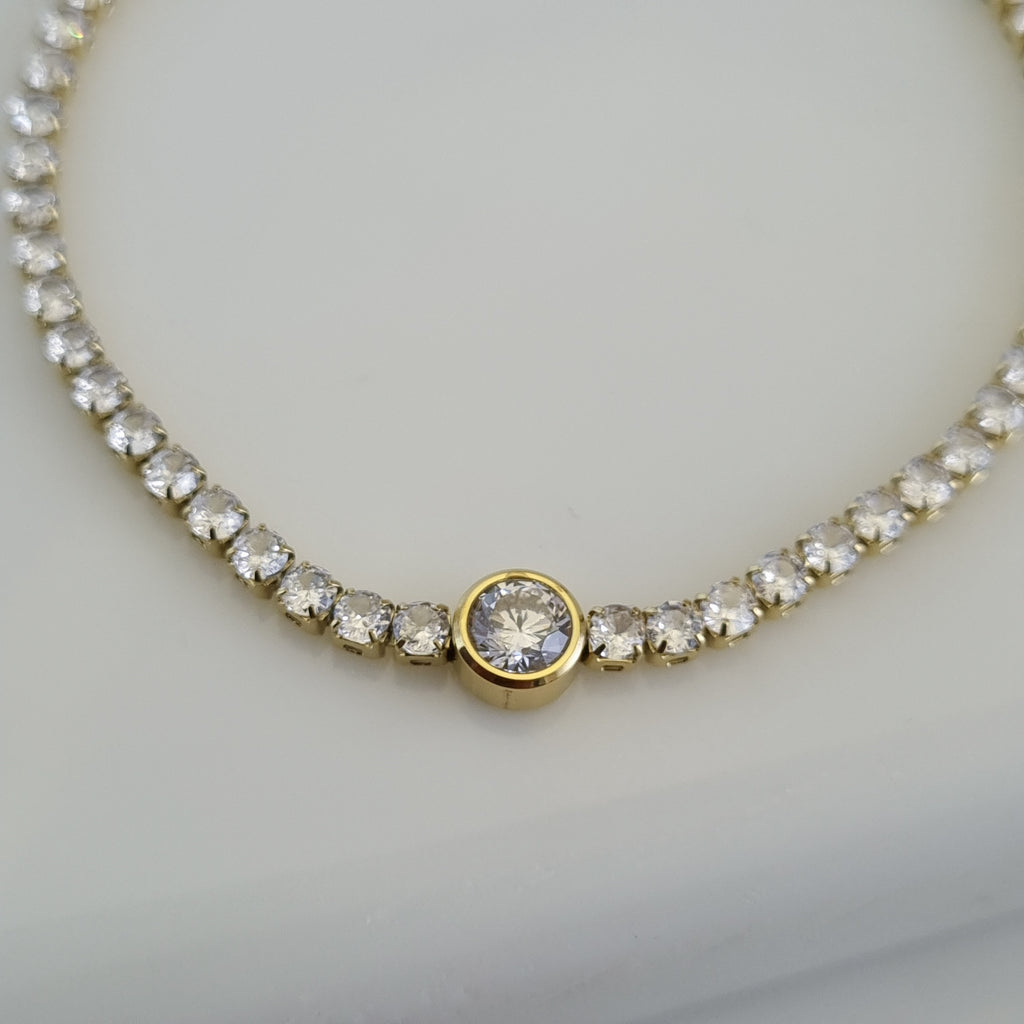 Treshire 18K gold plated tennis bracelet featuring a line of small crystals and a larger central bezel-set solitaire on a white surface.
