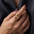 A woman with deep burgundy nails wearing an 18K gold-plated, size-adjustable triple star wrap ring on her index finger.