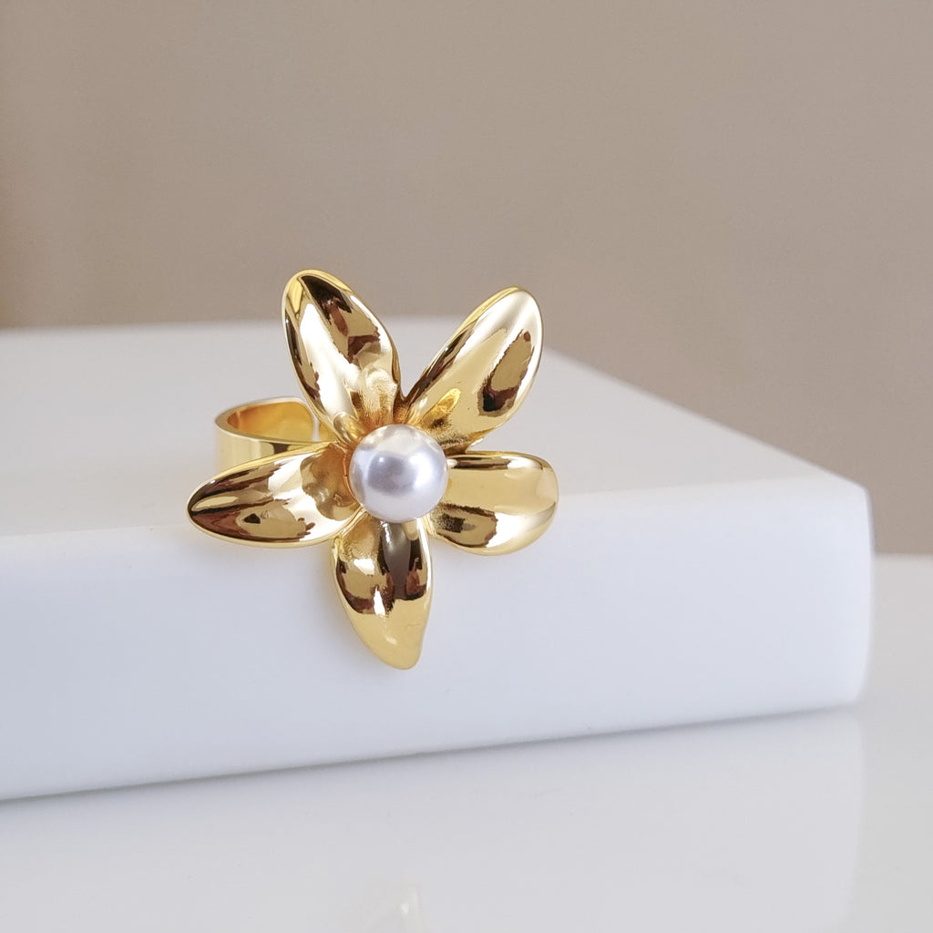 A high-polish 18K gold-plated, size-adjustable five-petal flower statement ring with a central pearl, displayed on a minimalist white surface.