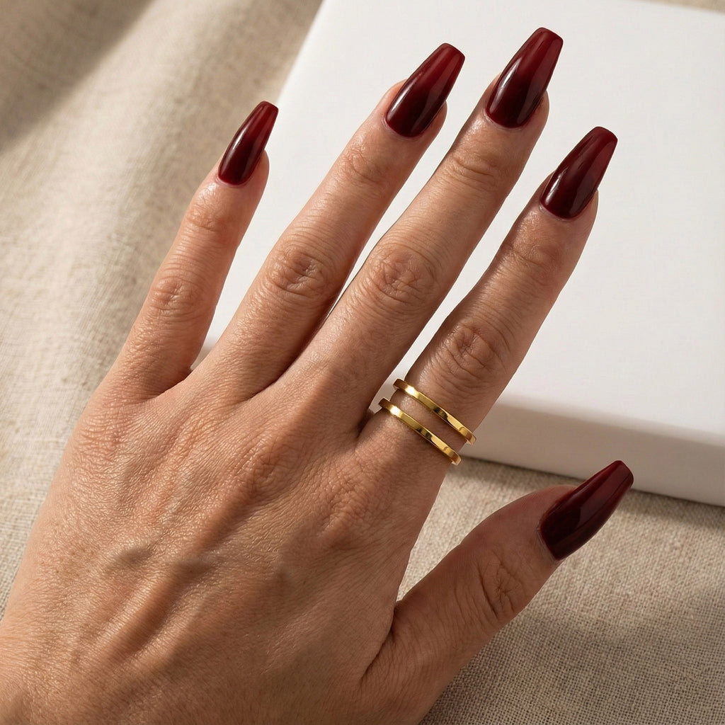 A woman with deep burgundy nails wearing an 18K gold-plated, size-adjustable parallel duo eternity band on her ring finger.