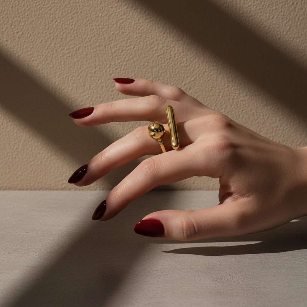 A woman with deep burgundy nails wearing an 18K gold-plated, size-adjustable abstract orb and bar statement ring on her middle finger.