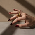 A woman with deep burgundy nails wearing an 18K gold-plated, size-adjustable abstract orb and bar statement ring on her middle finger.
