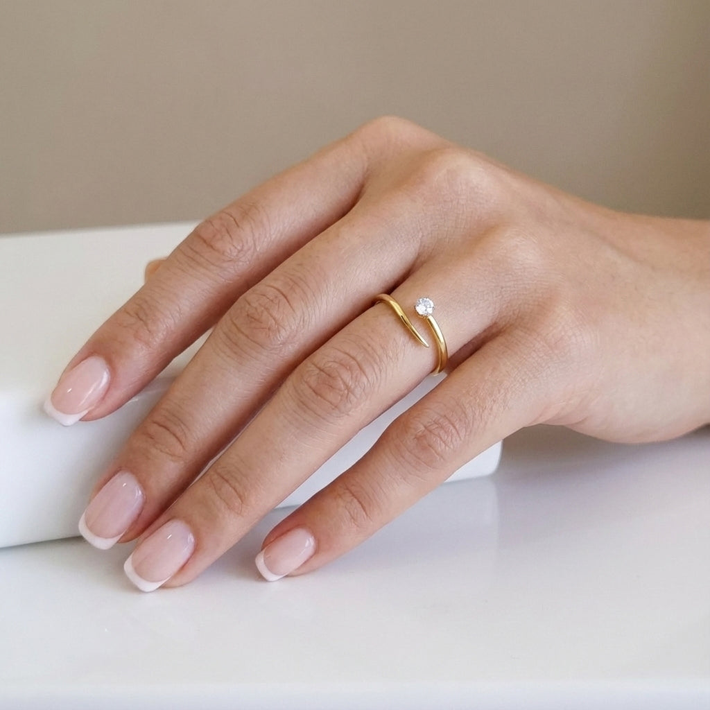 A high-polish 18K gold-plated, size-adjustable modern solitaire wrap ring with a single brilliant-cut crystal displayed on a woman's hand with a French manicure.