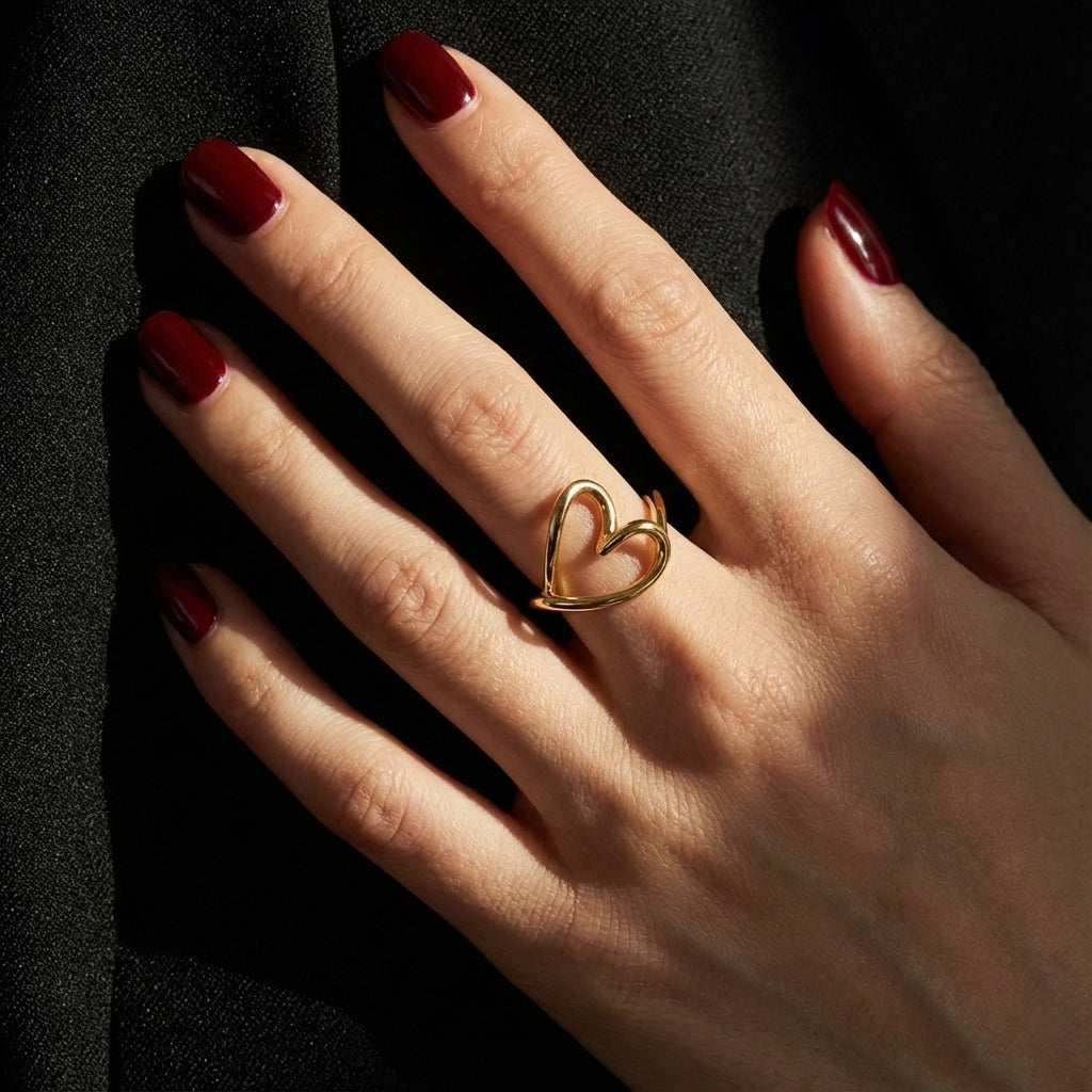 A woman with deep burgundy nails wearing an 18K gold-plated, size-adjustable organic open heart statement ring on her middle finger.