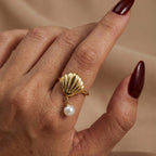 A woman with deep burgundy nails wearing an 18K gold-plated, size-adjustable scallop shell ring with a dangling pearl charm on her index finger.