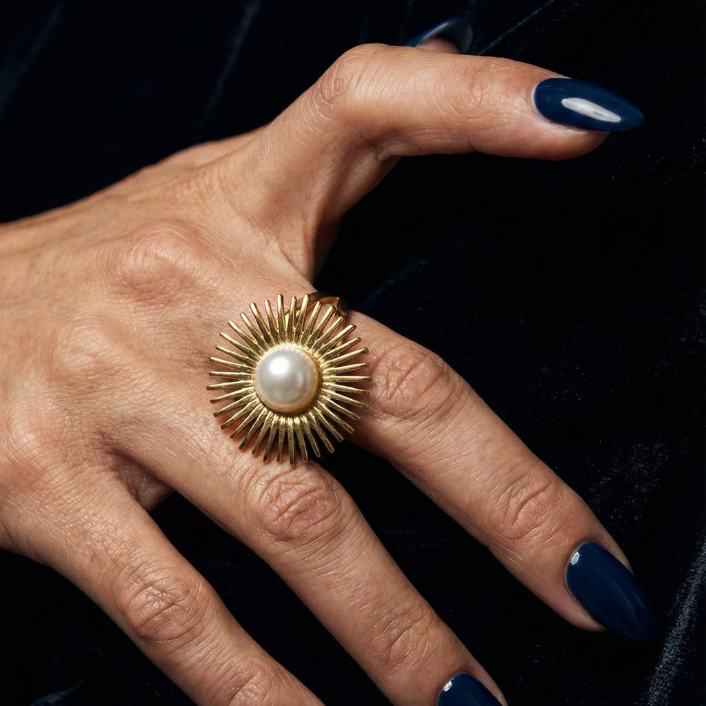 A woman with deep navy blue nails wearing a large 18K gold-plated, size-adjustable sunburst ring with a central pearl on her index finger.