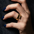 A woman with deep navy blue nails wearing a bold, 18K gold-plated, size-adjustable wide-band geometric wrap ring on her index finger.