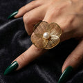 A woman with deep emerald green nails wearing a large 18K gold-plated, size-adjustable filigree flower statement ring with a central pearl on her index finger.