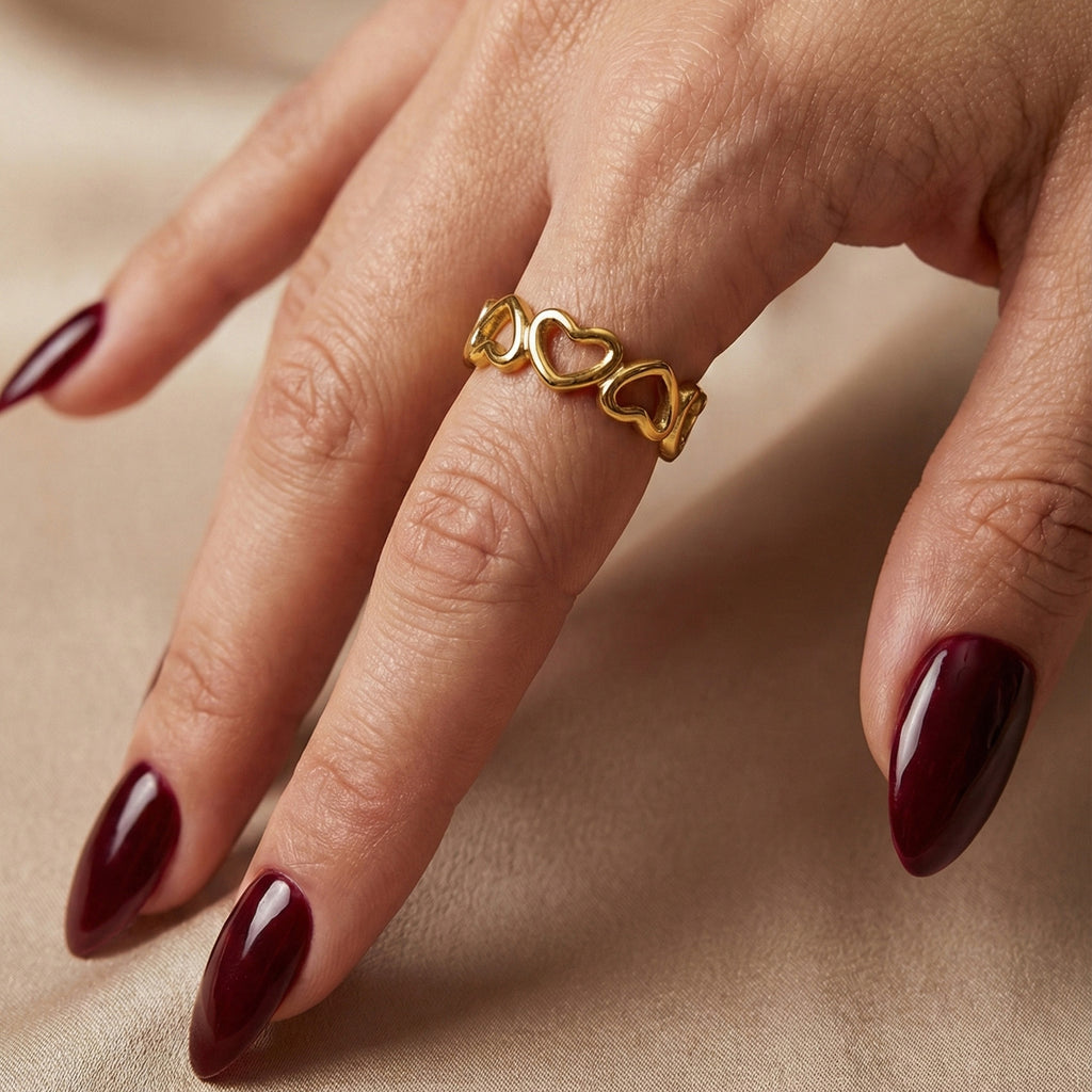 A woman with deep burgundy nails wearing an 18K gold-plated, size-adjustable infinite heart eternity band on her ring finger.