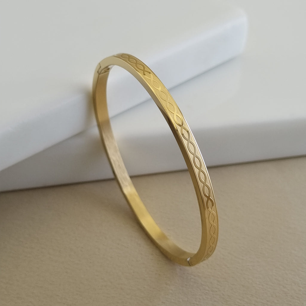 Treshire 18K gold plated Infinity Loop bangle with an etched interlocking loop pattern displayed on a white marble surface.