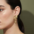 A woman with a sleek ponytail wearing large, 18K gold-plated sculptural earrings in a fluid, flame-like wing design that climbs the earlobe.